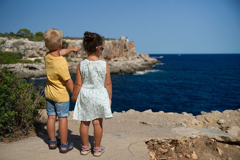 Vue de dos d'un petit garçon et d'une petite fille se tenant la main, au bord d'une falaise. L'enfant montre l'océan avec d'autres falaises au loin.