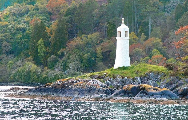 Un petit phare blanc sur une falaise en bord de mer, entouré de végétation.