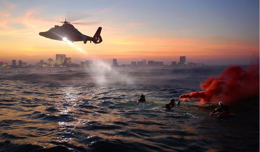 L'hélicoptère éclaire un groupe de personnes flottant dans la mer avec la fumée des fusées éclairantes, face à la ligne d'horizon de la ville à l'aube.