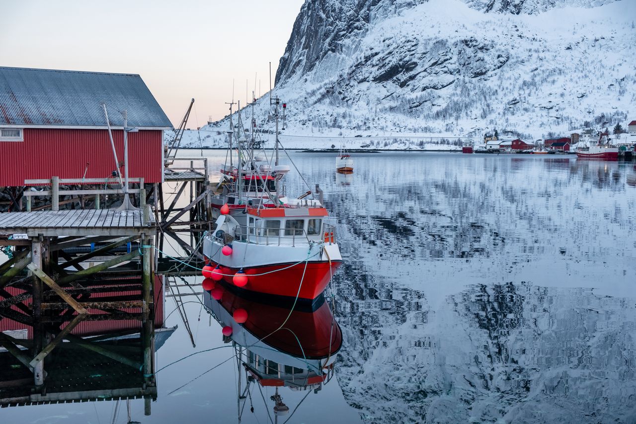 Bateau de pêche amarré dans les fjords des Lofoten, sous le soleil de minuit, près d’un village rouge.