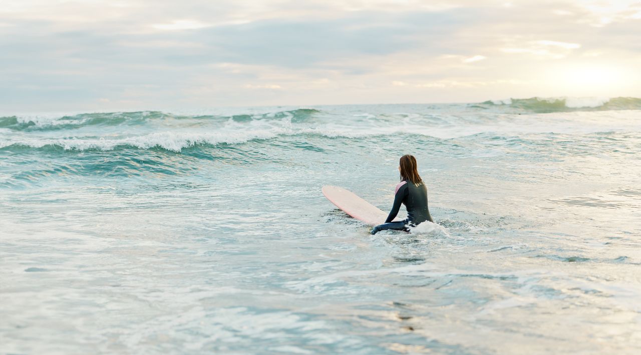 Surfeur préparant sa planche devant un surf camp simple, au bord de l’océan, lumière matinale.