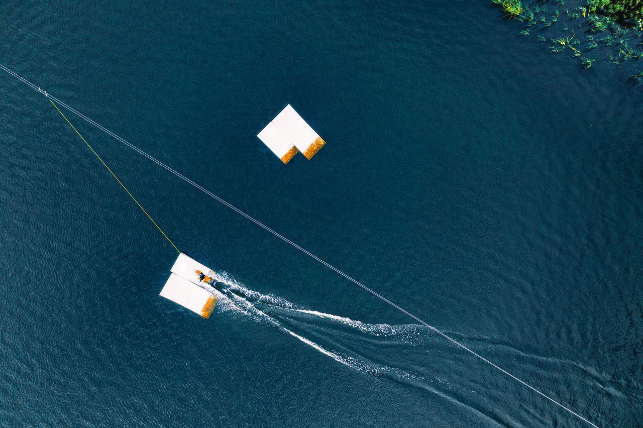 Wakeboardeur débutant en départ assis au cable park, câble tendu au-dessus d’une eau calme.